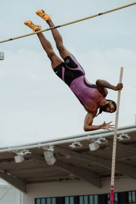 A pole vaulter crosses the bar in the flyover phase and holds the bar with one hand The athlete wears a purple and black one-piece suit and orange Puma spikes Pole vaulter crosses the bar during the competition Copyright: xspeedshotx xBeatricexBauerx