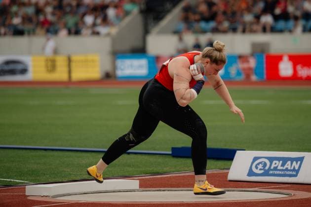 An athlete in a red jersey takes a shot put throw and holds the ball on her shoulder Spectators and advertising boards are visible in the background Athlete throwing a shot put in the stadium Copyright: xspeedshotx xBeatricexBauerx
