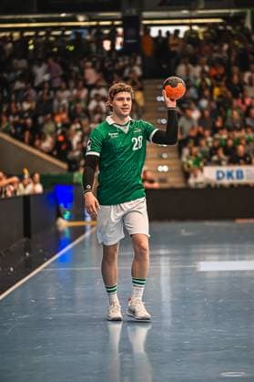 A Frisch Auf Göppingen player with back number 28 stands on the pitch and holds the handball up for a throw In the background you can see a well-filled spectator stand Frisch Auf Göppingen player 28 taking a throw Copyright: xspeedshotx xDorianfrickx
