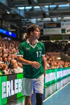 A Frisch Auf Göppingen player with jersey number 17 stands on the sideline and follows the game with concentration A well-filled spectator stand can be seen in the background Frisch Auf Göppingen player 17 watches the game Copyright: xspeedshotx xDorianfrickx