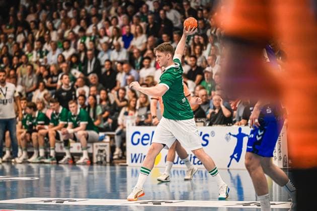 A Frisch Auf Göppingen player takes a throw with the ball in his raised hand A TVB Stuttgart player can be seen in the background, the stands are well filled Frisch Auf Göppingen players attempting a throw against TVB Stuttgart Copyright: xspeedshotx xDorianfrickx