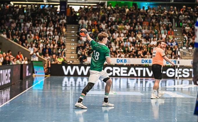 A Frisch Auf Göppingen player with the number 18 takes a shot while the opposing goalkeeper in the orange jersey takes up position The packed arena forms the backdrop for this scene Players 18 Frisch Auf Göppingen taking a shot in front of a full house Copyright: xspeedshotx xDorianfrickx