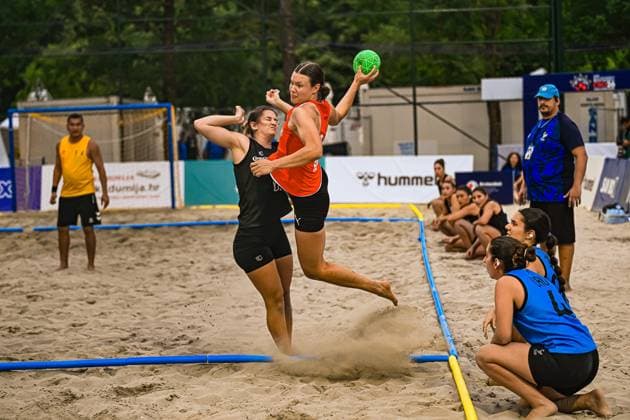 Eine Spielerin im orangefarbenen Trikot springt mit dem Ball zum Wurf ab, während eine Gegenspielerin in Schwarz sie bedrängt. Im Sand wirbelt Staub auf, am Spielfeldrand beobachten weitere Spielerinnen in Blau die Szene. IHF Beach Handball World Championship, WM, Weltmeisterschaft 2026, 23.06.2026, Zagreb, Jarun See *** A player in an orange jersey jumps up to shoot the ball while an opponent in black pressures her. Dust swirls up in the sand; on the sidelines, other players in blue watch the scene. 2026 IHF Beach Handball World Championship, June 23, 2026, Zagreb, Lake Jarun,detected_source_language Copyright: xspeedshotx xDorianfrickx