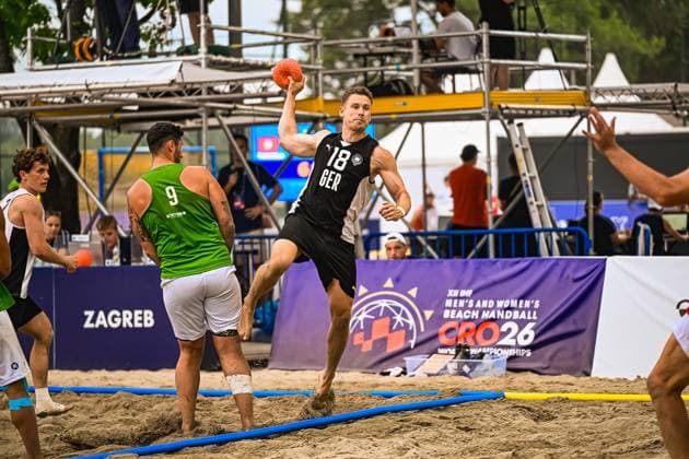Robin John ( 18; Deutschland Beachhandball Nationalmannschaft) springt zum Wurf und hält den Ball hoch über dem Kopf, während ein Gegenspieler mit 9 im grünen Trikot den Abschluss zu blocken versucht. Die Szene findet im Sand statt, im Hintergrund ist das Banner der IHF Beach Handball World Championships CRO26 in Zagreb zu sehen. IHF Beach Handball World Championship, WM, Weltmeisterschaft 2026, 23.06.2026, Zagreb, Jarun See *** Robin John, 18, of the German national beach handball team, jumps to shoot and holds the ball high above his head, while an opponent wearing a green jersey with the number 9 tries to block the shot. The scene takes place on the sand, and in the background is the banner for the IHF Beach Handball World Championships CRO26 in Zagreb. IHF Beach Handball World Championship 2026, June 23, 2026, Zagreb, Lake Jarun,detected_source_language Copyright: xspeedshotx xDorianfrickx