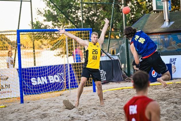Moritz Ebert ( 26; Deutschland Beachhandball Nationalmannschaft) streckt sich im Tor und versucht einen Wurf des Gegners abzuwehren, während der Ball bereits hoch in der Luft ist. Ein Angreifer im blauen Trikot springt zum Wurf, im Vordergrund ist ein weiterer Spieler im roten Trikot zu sehen. IHF Beach Handball World Championship, WM, Weltmeisterschaft 2026, 23.06.2026, Zagreb, Jarun See *** Moritz Ebert, 26, of the German national beach handball team, stretches toward the goal and tries to block an opponent’s shot while the ball is already high in the air. An attacker in a blue jersey jumps to take a shot, and another player in a red jersey can be seen in the foreground. 2026 IHF Beach Handball World Championship, June 23, 2026, Zagreb, Lake Jarun Copyright: xspeedshotx xDorianfrickx