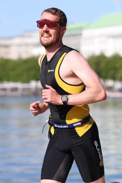 Ein Triathlet in schwarzem und gelbem Neoprenanzug mit Sportsonnenbrille läuft entlang der Binnenalster in Hamburg. Im Hintergrund sind die Uferpromenaden und Gebäude des Jungfernstiegs sichtbar. Triathlet in Wettkampfausrüstung beim Hamburg Triathlon an der Binnenalster *** A triathlete in a black and yellow wetsuit and athletic sunglasses is running along the Binnenalster in Hamburg. In the background, the waterfront promenades and buildings of the Jungfernstieg are visible. Triathlete in competition gear at the Hamburg Triathlon on the Binnenalster Copyright: xspeedshotx xSarahxWolfx
