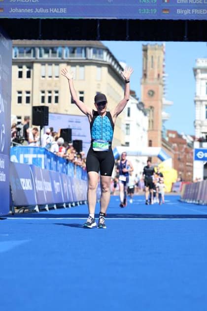 Eine Triathletin überquert die Ziellinie am Jungfernstieg in Hamburg mit erhobenen Armen in Jubelpose. Sie trägt einen grünen Helm, blaues Triathlon-Top und schwarze Shorts mit Startnummer. Im Hintergrund sind historische Hamburger Gebäude und eine Anzeigetafel mit Zeiten sichtbar. Triathletin im Zieleinlauf mit Jubelpose am Jungfernstieg Hamburg *** A female triathlete crosses the finish line at Jungfernstieg in Hamburg with her arms raised in a celebratory pose. She is wearing a green helmet, a blue triathlon top, and black shorts with her race number. In the background are historic Hamburg buildings and a scoreboard displaying times. Triathlete crossing the finish line in a celebratory pose at Jungfernstieg, Hamburg Copyright: xspeedshotx xSarahxWolfx