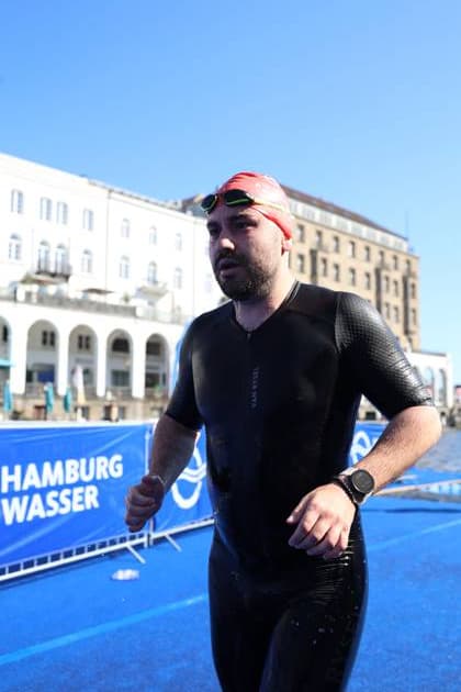 Ein Läufer in schwarzem Neoprenanzug und roter Schwimmkappe absolviert die Laufstrecke des Triathlons am Jungfernstieg in Hamburg. Im Hintergrund sind die klassischen Fassaden der Alsterarkaden und das klare Wasser der Binnenalster zu sehen. Triathlet in der Laufstrecke am Jungfernstieg, Hamburg *** A runner in a black wetsuit and red swim cap completes the running leg of the triathlon on Jungfernstieg in Hamburg. In the background are the classic facades of the Alsterarkaden and the clear waters of the Binnenalster. Triathlete on the running course at Jungfernstieg, Hamburg Copyright: xspeedshotx xSarahxWolfx