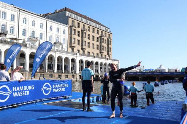 Triathleten in Neoprenanzügen nach dem Schwimmen am Jungfernstieg. Im Hintergrund die klassische Hamburger Architektur mit Alsterarkaden und Binnenalster-Ufer, blauer Himmel und Veranstaltungsinfrastruktur mit HAMBURG WASSER und Suzuki-Sponsoring. Triathleten beim Schwimmstart an der Binnenalster, Hamburg *** Triathletes in wetsuits after the swim at Jungfernstieg. In the background: classic Hamburg architecture featuring the Alster Arcades and the Binnenalster waterfront, a blue sky, and event infrastructure with HAMBURG WASSER and Suzuki sponsorship. Triathletes at the start of the swim at the Binnenalster, Hamburg Copyright: xspeedshotx xSarahxWolfx