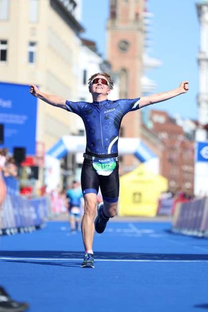 Ein Triathlet in dunkelblauem ARRIBA-Rennanzug und grünem Helm überquert die Zielline am Jungfernstieg in Hamburg mit ausgebreiteten Armen in Jubelpose. Im Hintergrund sind historische Hamburger Gebäude und eine rote Kirchturmspitze sichtbar, Oscar Seydel-Winter ( 3192, Sprintdistanz) Triathlet überquert Zielline mit Jubelgeste am Jungfernstieg Hamburg, Oscar Seydel-Winter 3192, *** A triathlete in a dark blue ARRIBA racing suit and green helmet crosses the finish line at Jungfernstieg in Hamburg with his arms outstretched in a celebratory pose. In the background, historic Hamburg buildings and a red church spire are visible. Oscar Seydel Winter 3192, A sprint-distance triathlete crosses the finish line with a celebratory gesture at Jungfernstieg in Hamburg, Oscar Seydel Winter 3192, Copyright: xspeedshotx xSarahxWolfx