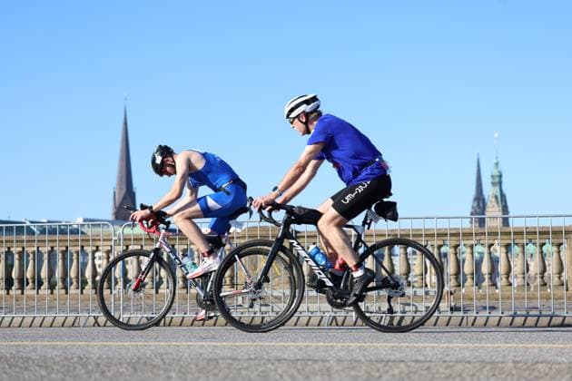 Zwei Athleten absolvieren die Radeinheit beim Triathlon am Jungfernstieg in Hamburg, im Hintergrund sind die Türme der Hamburger Innenstadt zu sehen. Der vordere Fahrer trägt einen blauen Trisuit mit der Startnummer 1439, der hintere Athlet ein blaues Trikot mit schwarzer Radhose. Zwei Triathleten auf der Radstrecke am Jungfernstieg Hamburg *** Two athletes are completing the cycling leg of the triathlon at Jungfernstieg in Hamburg, with the towers of downtown Hamburg visible in the background. The rider in front is wearing a blue trisuit with race number 1439, while the athlete behind is wearing a blue jersey and black cycling shorts. Two triathletes on the bike course at Jungfernstieg in Hamburg Copyright: xspeedshotx xSarahxWolfx