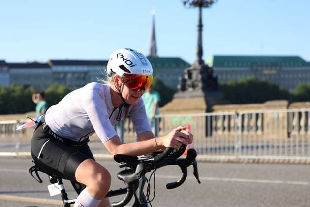 Eine Triathletin mit der Startnummer 1195 fährt auf einem Canyon-Rennrad auf der Radstrecke am Jungfernstieg in Hamburg. Sie trägt einen weißen EKOI-Helm mit roten Sportgläsern und lächelt in die Kamera. Triathletin ( 1195) auf der Radstrecke am Jungfernstieg Hamburg *** A triathlete with bib number 1195 is riding a Canyon road bike on the cycling course at Jungfernstieg in Hamburg. She is wearing a white EKOI helmet with red sports glasses and is smiling at the camera. Triathlete 1195 on the cycling course at Jungfernstieg, Hamburg Copyright: xspeedshotx xSarahxWolfx