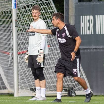 Ein Torwart des FC St. Pauli steht vor dem Tor, während ein Trainer in schwarzem Puma-Shirt mit den Initialen MR ihm Anweisungen gibt und eine Positionierung demonstriert. Die Szene findet auf dem Trainingsgelände des FC St. Pauli in Hamburg statt. Torwarttraining beim FC St. Pauli im Trainingszentrum Hamburg *** An FC St. Pauli goalkeeper stands in front of the goal while a coach wearing a black Puma shirt with the initials MR gives him instructions and demonstrates proper positioning. The scene takes place at the FC St. Pauli training facility in Hamburg. Goalkeeper training at FC St. Pauli s training center in Hamburg Copyright: xAndreasxTolksdorfx