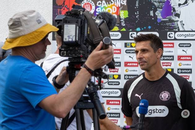 Ein Verantwortlicher des FC St. Pauli gibt vor der Sponsorenwand des Trainingszentrums in Hamburg ein Interview mit dem NDR. Ein Kameramann filmt die Szene, im Hintergrund ist das Saisonmotto Causing Good 2026 27 zu sehen. FC St. Pauli-Verantwortlicher im NDR-Interview am Trainingszentrum *** An FC St. Pauli official gives an interview to NDR in front of the sponsor wall at the training center in Hamburg. A cameraman films the scene; in the background, the season’s motto, Causing Good 2026–27, is visible. FC St. Pauli official in an NDR interview at the training center Copyright: xspeedshotx xAndreasxTolksdorfx