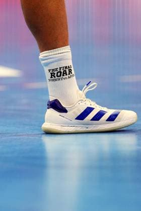 Nahaufnahme eines weißen Adidas Adizero Fastcourt Handballschuhs auf dem blauen Hallenboden der SAP Arena. Die weiße Socke trägt den Schriftzug The Final Roar – Johnny vs. Apfel . Detail: Adidas Adizero Fastcourt mit The Final Roar -Socke in der SAP Arena *** Close-up of a white Adidas Adizero Fastcourt handball shoe on the blue indoor court floor of the SAP Arena. The white sock features the lettering The Final Roar – Johnny vs. Apfel. Detail of the Adidas Adizero Fastcourt with The Final Roar sock at the SAP Arena Copyright: xspeedshotx xLukasxTrunkx