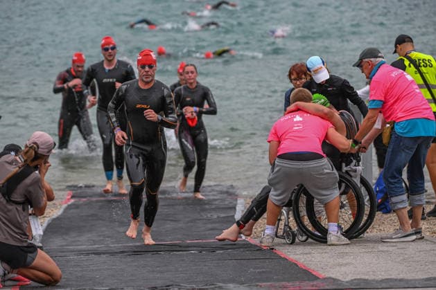 TRIATHLON: Stepniak And Bartlett Secure Elite Victories At IRONMAN 70.3 Krakow KRAKOW, POLAND - AUGUST 02: Volunteers in pink shirts embrace and assist a paratriathlete into a racing wheelchair on the shoreline as other age-group competitors run out of the water during the swim-to-bike transition stage of the Enea IRONMAN 70.3 Krakow triathlon at the Zakrzowek Reservoir in Krakow, Lesser Poland Voivodeship, Poland, on August 02, 2026. Over 2,100 registered athletes completed the opening 1.9-kilometer open-water swim phase inside the flooded limestone quarry before rushing toward the first transition zone (T1) to begin the 90-kilometer cycling segment. Krakow Poland  Copyright: xArturxWidakx originalFilename:widak-triathlo260802_npZ78.jpg