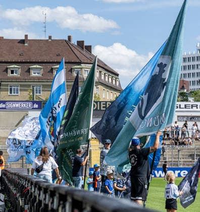The flag wavers in front of the standing-room-only section before kickoff: GER, TSV 1860 Munich vs. TSV Landsberg 1882, soccer, Bavarian Regional League, Matchday 6, 2026–2027 season, August 30, 2026. DFL and DFB regulations prohibit any use of photographs as image sequences and or quasi-video. Photo: HMB Media, Heike Feiner Copyright: HMBxMedia HeikexFeiner