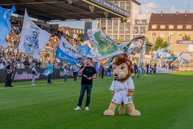 Sebastian Schaech, stadium announcer for TSV 1860 Munich, and the mascot Sechzgerl in front of the fan section; in the background, the flag wavers in front of the GER standing section, TSV 1860 Munich vs. TSV Schwaben Augsburg, soccer, Bavarian Regional League, Matchday 8, 2026–2027 season, September 11, 2026 DFL and DFB regulations prohibit any use of photographs as image sequences and or quasi-video. Photo: HMB Media, Heike Feiner Copyright: HMBxMedia HeikexFeiner