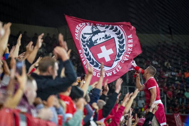 Soccer UEFA Nations League 2026–2027, Matchday 3: Switzerland vs. Slovenia at Thermoplan Arena Lucerne. Swiss fans in the stands celebrate during the international match; a chant leader with a megaphone stands in front of a large red flag of the Swiss Nati Supporters featuring the Swiss cross. Spectators, fans, fan culture, fan section, stands, flag, fan flag, Swiss cross, megaphone, cheerleader, atmosphere, support, Swiss national team, Nati, soccer, UEFA Nations League Copyright: xJOERANxSTEINSIEKx