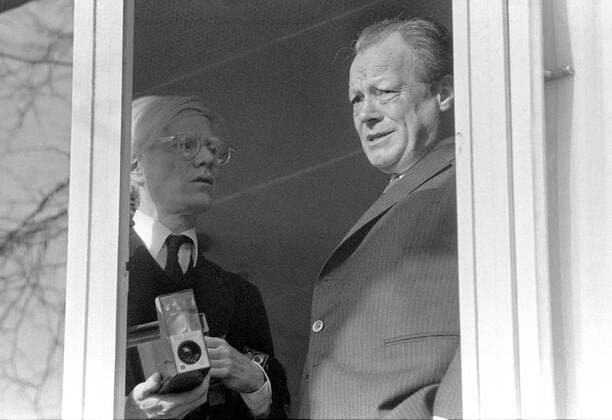 Bundeskanzler Willy Brandt (re., GER SPD) steht mit dem Künstler Andy Warhol (USA) am Fenster