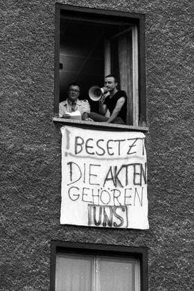Bürgerrechtler Rolf Henrich (li., GER Neues Forum) und Protestler mit Megaphon am geöffneten Fenster über einem Transparent während der Besetzung des HAUPTQUARTIERS DES MINISTERIUMS FÜR STAATSSICHERHEIT DER DDR in Berlin-Lichtenberg