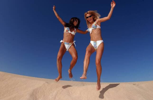 Zwei Freundinnen in Bikinis springen jubelnd von einer Sanddüne
