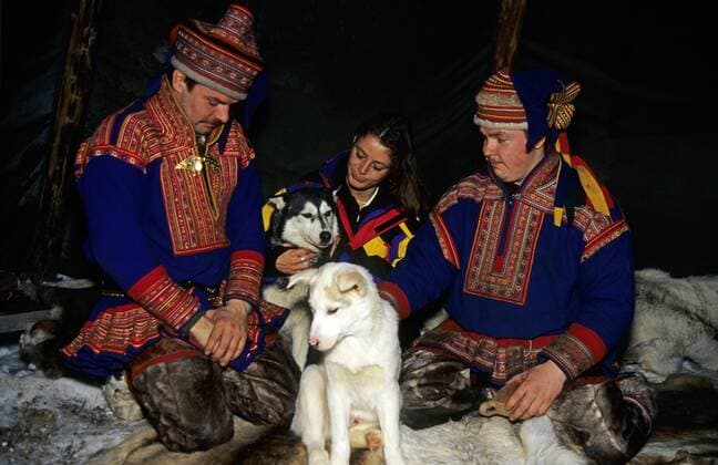Zwei Männer in den Trachten der Sami und eine Frau streicheln ihre Huskys im Nomadenzelt auf einer Schlittenfahrt - Muonio Lappland
