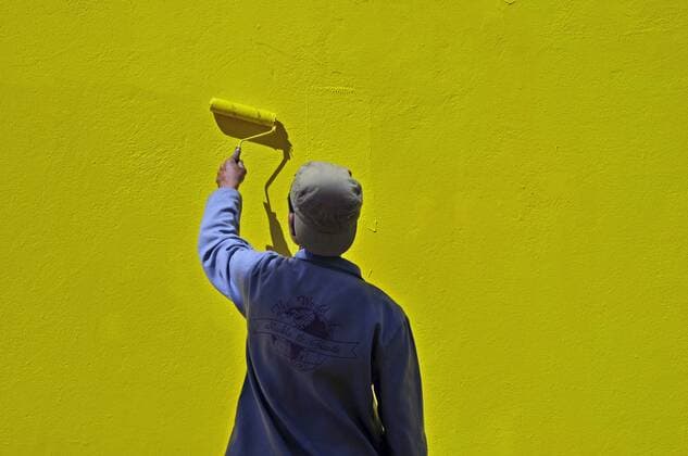 junger Mann mit Baseballkappe streicht mit Farbrolle Hauswand gelb, Suedafrika, Kapstadt young man wearing a baseball cap is painting a house fassade yellow using a paint roller, South Africa, Bo-Kaap, Capetown BLWS241426