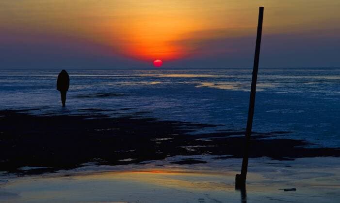 Silhouette einer Frau, die bei Ebbe im Watt stehend auf die Nordsee hinausschaut und den Sonnenuntergang betrachtet, Deutschland, Niedersachsen, Landkreis Cuxhaven, Cuxhaven silhouette of a woman standing at the wadden sea at low tide looking out at the North sea and watching the sunset, Germany, Lower Saxony, Landkreis Cuxhaven, Cuxhaven BLWS250586