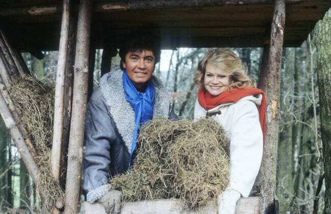 Rex Gildo mit Ehefrau Marion 12 84 geue leger Jacke Stiefel draußen Futterkrippe Futter Heu Stand draußen Wald Baum Natur kniend halb quer