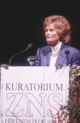 Hannelore Kohl (Ehefrau des Bundeskanzlers) 11 86 - Frau Gesellschaft Politik dunkelhaarig kurzhaarig Locken Schmuck Ohrring Kette seriös Jacke Blazer blau Bluse weiss stehend Saal Halle dunkel Pult Rednerpult Schrift Kuratorium für Unfallverletzte mit Schäden des Zentralnervensystems ZNS halb hoch sprechend redend zur Seite schauend Halbprofil Mikrophon Glas Weinglas Getränk Wasser