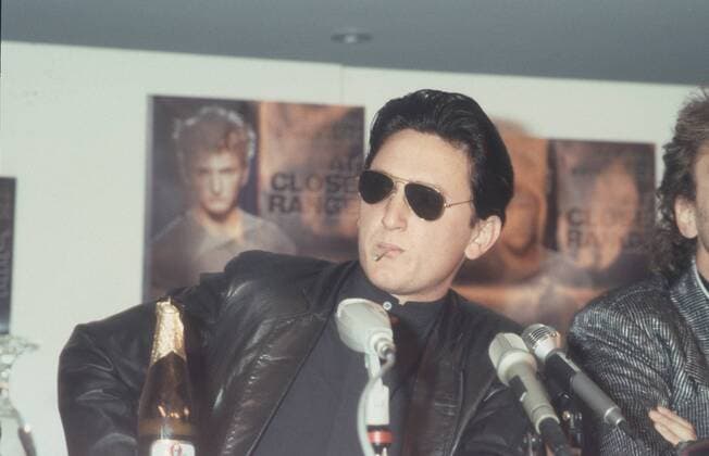 Sean Penn (Schauspieler) 02 86 - Sean Penn im Februar 1986 während der Berliner Filmfestspiele Berlin Filmfestival Film Kino Mann USA Amerikaner amerikanischer Amerika TV Fernsehen Schauspiel Regisseur Regie american actor director Bühne Podium Mikrofon Pilsener Pils Bier Bierflasche Plakat Filmplakat at close range auf kurze Distanz PK Presskonferenz Pressegespräch quer halb sitzend Sonnenbrille dunkelhaarig rauchen rauchend Zigarette cool