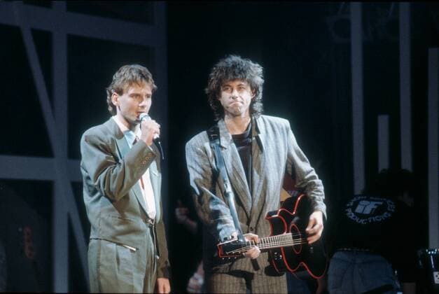 Bob Geldof (re.) mit Peter Illmann 11 86 vZ Bob Geldof (re.) mit Peter Illmann am 21.11.1986 in Peters Pop Show , ZDF - Sendedatum 6.12.1986 Irland Ire irischer Mann Popmusik Pop Rockmusik Rock Musik Musiker Instrument Texter Komponist TV Fernsehen Sänger Musiksendung Deutschland deutsch Deutscher Mann Moderator Mikrofon neutral Moderation irish man musician singer german tv-host actor songwriter presenter Bühne Gitarre haltend spielend lächelnd Anzug dunkelhaarig lockig Locken halb quer