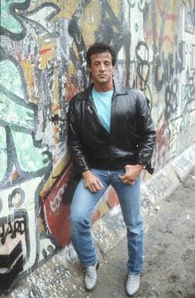 Sylvester Stallone (Schauspieler) 06 88 - Sylvester Stallone im Juni 1988 an der Berliner Mauer Promotiontour Promotion Filmstart Deutschland Rambo III Berlin Grenze DDR Ostberlin Ost West Sly Mann Kino Film Schauspiel Schauspieler USA Amerika amerikanischer Amerikaner american actor germany US wall hoch ganz draußen stehend angelehnt anlehnend freistellbar Graffiti