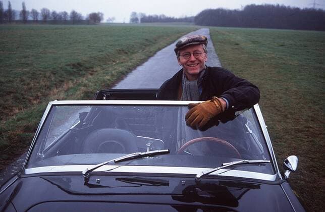 Joachim-Hermann Luger mit Austin Healey 03 97 teu Mann TV Fernsehen Schauspiel Serie grauhaarig Mütze leger Lederjacke Lederweste Brille draußen sitzend Auto aufstützend Steuerrad haltend Straße Landschaft Feld Wiese halb quer lächelnd