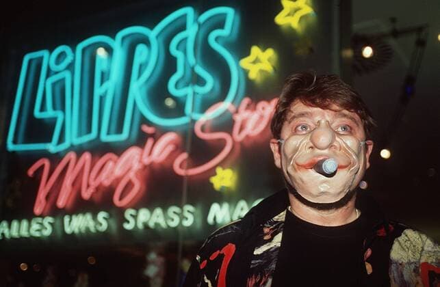 Jürgen von der Lippe vor seinem Laden Lippes Magic Store 05 98 pp Mann TV Fernsehen Schauspiel Moderation Show Komik Kabarett Comedy Musik Gesang dunkelhaarig leger T-Shirt Hemd Bart stehend Schild Logo Geschäft halb quer Maske tragend