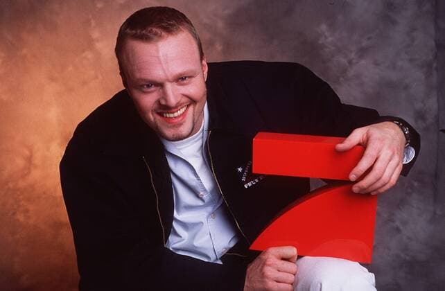 Stefan Raab (TV-Moderator, Musiker) 01 99 her Mann TV Moderation Musik VIVA Unterhaltung Produktion Gesang Pop TV Fernsehen Tv total Logo Schild haltend PRO 7 ProSieben lächelnd kurzhaarig leger Shirt aufstützend sitzend halb quer