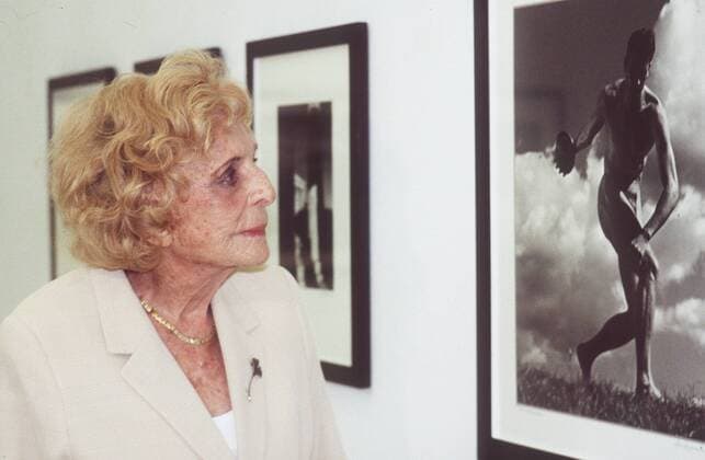 Leni Riefenstahl (Fotografin) 05 00 ker Frau Fotografie Kunst Handwerk Künstlerin umstritten Nazi-Zeit dunkelhaarig blondiert blond Locken Schmuck Kette seriös Jacke Blazer beige kariert Top weiß stehend vor Wand weiß mit Schwarzweiß-Bildern Bilder schwarz-weiß SW (Ausstellung mit Photographien Photos der Olympiade 1936 in Berlin) Galerie Raum Saal Halle Ausstellungshalle Ausstellung halb quer nachdenklich lächelnd ihre Werke betrachtend Fototermin Leni Riefenstahl am 04.05.2000 in Berlin