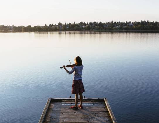 A ten year old girl playing the violin at dawn on a wooden dock. 