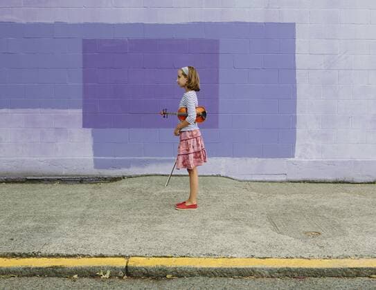 A ten year old girl holding a violin under her arm and a bow in her hand. Against a purple background. 