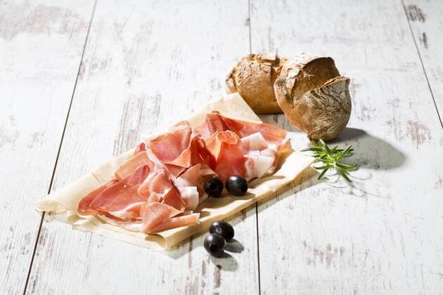 Parma ham on greaseproof paper, rolls and black olives on bright wood  MAEF009791