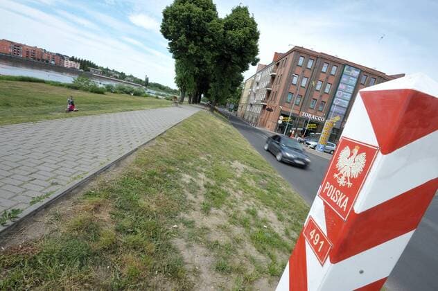 Zwischen Frankfurt Oder und Slubice liegt die Grenze zwischen Deutschland und Polen. Auf polnischer Seite markieren Grenzpfähle in den Farben Rot und Weiß mit dem Landeswappen und der Aufschrift Polska den Grenzübertritt. Links sind die Häuser von Frankfurt Oder zu sehen, die Oder als Grenzfluss. Auf dem als Hochwasserschutz aufgeschütteten Damm befindet sich ein geplasteter Weg. Grenzübergang Deutschland-Polen