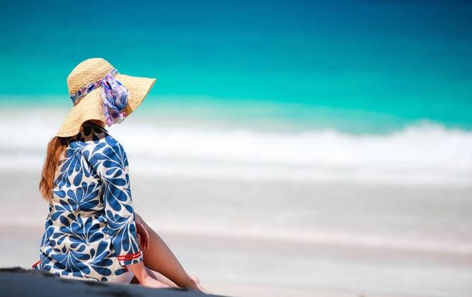eine Frau mit Sonnenhut sitzt am Sandstrand, Rueckansicht woman with sun hat sitting on sandy beach, rear view BLWS269835