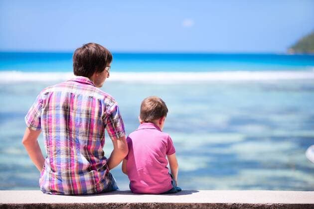 Vater sitzt mit seinem Sohn auf einer Mauer und blickt auf das Meer, Rueckansicht father with his son sitting on wall and viewing to the sea, rear view BLWS269841