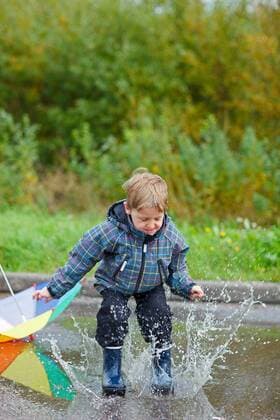 kleiner Junge mit Gummistiefel springt in eine Pfuetze little boy jumping with rain boots in a puddle BLWS272234