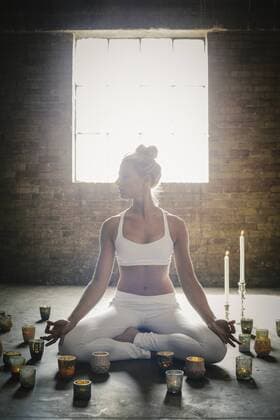 A blonde woman in a white crop top and leggings, sitting on the floor surrounded by candles, doing yoga. A blonde woman in a white crop top and leggings, sitting on the floor surrounded by candles, doing yoga.  16701