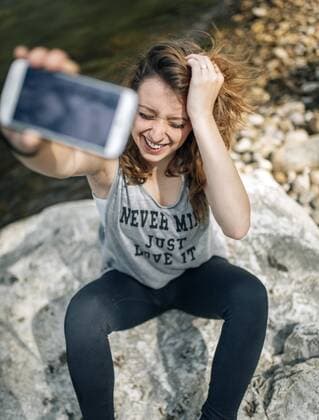 Portrait of a laughing young woman sitting on a rock taking selfie with a smartphone model released Symbolfoto  MGOF000217