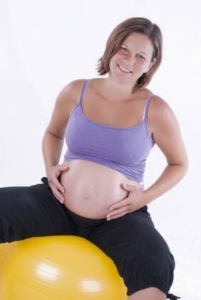 schwangere junge Frau sitzt auf Gymnastikball und haelt ihren runden Bauch pregnant young woman sitting on a gym ball holding her round belly BLWS242759