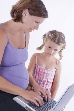 schwangere junge Frau arbeitet am Laptop auf ihrem Schoss, kleine Tochter steht daneben und schaut zu pregnant young woman working on the laptop in her lap, young daughter standing beside watching BLWS242760
