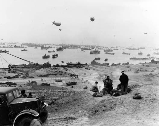 View of the east end of the Mulberry A artificial harbor off Omaha Beach, showing the Gooseberry breakwater of sunken ships and unloading operations at the shoreline. Among the vessels on the beach are USS LCT-149 at left, and USS LCT-195 just to the right of center. Photographed by the Office of Strategic Services. It was received by the Naval Photographic Science Laboratory on November 21, 1944, but actually taken in June of that year. Note beached seamen with their gear in the center foreground, and Royal Navy vehicle at the extreme lower left.  WAX2016060140 U.S.xNavy